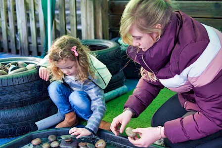 An early years practitioner with a young girl. She is teaching the young girl about shapes and textures using stones from an outdoor pond.