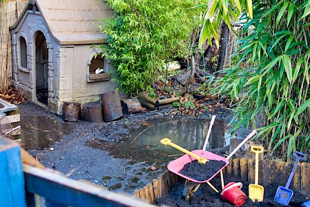 The nursery garden. A playhouse sits next to a large puddle which the children can jump in. There is a wheelbarrow in front filled with mud. It looks like children have been making castles in the mud with buckets and spades.